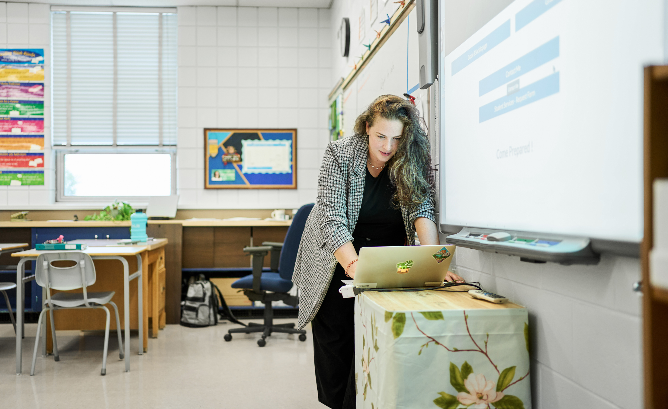 School teacher preparing for a lesson using a screen projector and a laptop while working at a desk in her classroom (credit: iStock / AJ_Watt)